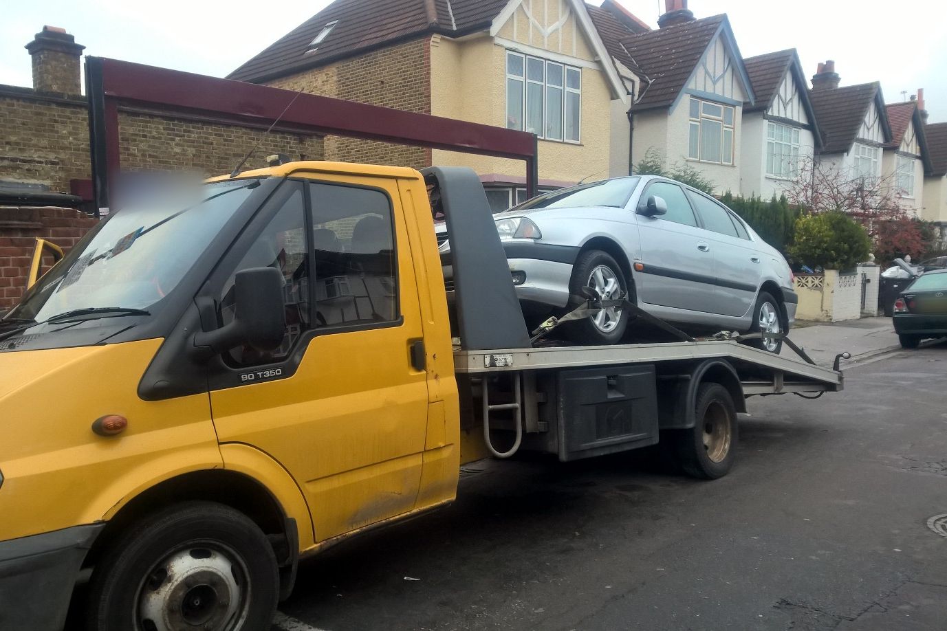 Silver hatchback strapped onto a tilt-bed recovery truck on a UK residential street